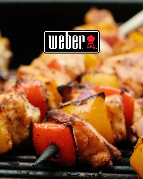 Nahaufnahme von gegrillten Hähnchen- und Gemüsespießen auf einem Grill. Die Spieße bestehen aus Hähnchenstücken mit roten und gelben Paprikaschoten sowie roten Zwiebeln. Das Weber-Logo ist prominent oben in der Bildmitte zu sehen.