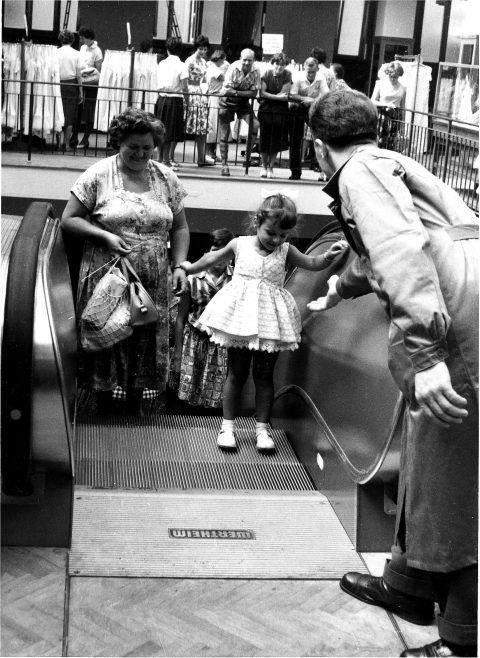 Ein Schwarzweißfoto zeigt ein junges Mädchen im Kleid, das mit Hilfe eines Rolltreppenwärters eine Rolltreppe betritt. Ihre Mutter steht lächelnd daneben. Hinter einem Geländer beobachten Zuschauer das Mädchen. Die Szenerie scheint ein geschäftiges Kaufhaus zu sein.