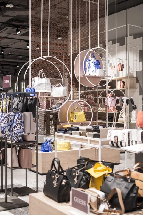 In einem modernen Ladeninneren wird eine Auswahl an Handtaschen in verschiedenen Farben und Stilen präsentiert. Die Taschen werden auf runden und rechteckigen Ständern vor einem Hintergrund aus geometrischen, offenen Regalgittern und warmer Beleuchtung präsentiert. Auf einem Schild steht „GUESS“.