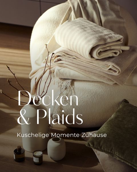 Ein gemütlicher Stuhl mit gefalteten beigen und gestreiften Decken, ein grünes Kissen, weiße Vasen, Zweige und Kerzen auf einem Holzboden. Der deutsche Text lautet: "Decken & Plaids Kuschelige Momente Zuhause.