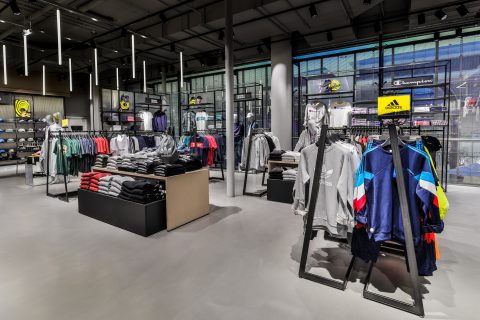 Ein moderner Sportbekleidungsladen verfügt über ordentlich angeordnete Kleiderständer mit verschiedenen Hemden, Jacken und Kapuzenpullovern. Markenlogos wie Adidas und Champion sind sichtbar. Der Laden verfügt über eine elegante Beleuchtung und ein großzügiges Layout.