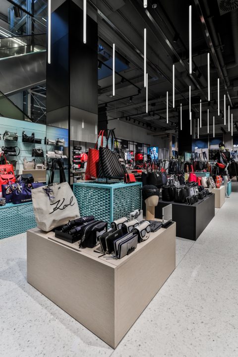 Modernes Ladeninterieur mit elegantem Design und lebendiger Beleuchtung. Auf Holzständern werden verschiedene Handtaschen und Accessoires ausgestellt. Hängende Deckenleuchten sorgen für Eleganz vor einem Hintergrund aus Glas und Metall.