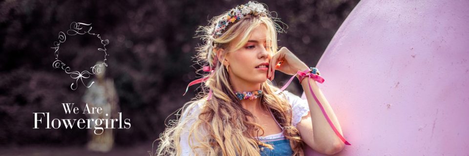 Eine Frau mit langen blonden Haaren und einer Blumenkrone lehnt an einem großen rosa Objekt. Sie trägt ein Dirndlkleid mit einer Blumenkette und Bänderarmbändern. Der Text lautet „We Are Flowergirls“ mit einem dazugehörigen kreisförmigen Blumenlogo.