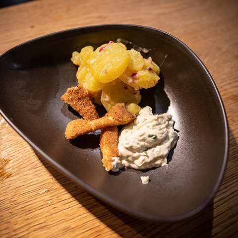 Ein dunkler, dreieckiger Teller auf einer Holzoberfläche, präsentiert eine Portion panierte Garnelen, einen Kartoffelsalat mit Kartoffelscheiben und roten Zwiebeln sowie einen Klecks cremige Kräutersauce.