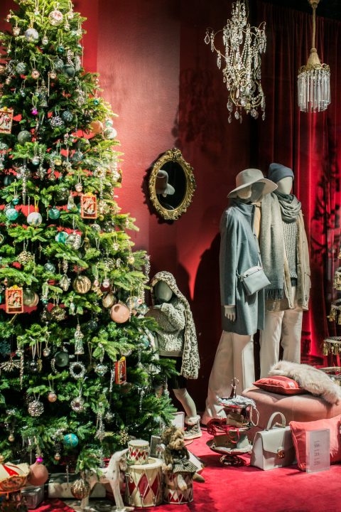 In einem festlichen Schaufenster ist ein geschmückter Weihnachtsbaum mit Ornamenten zu sehen. Daneben sind zwei Schaufensterpuppen in Winterkleidung zu sehen. Zur Dekoration gehören ein Kronleuchter, ein Spiegel und verschiedene Geschenkboxen, alles vor leuchtend roten Vorhängen.