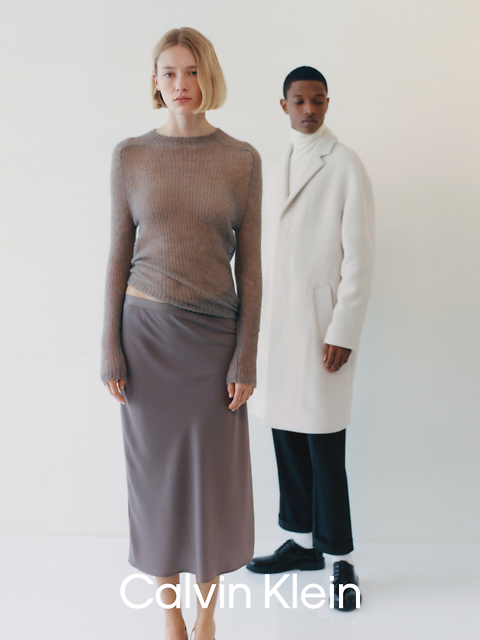 Eine Frau in einem transparenten Langarmshirt und einem langen Rock steht vor einem Mann, der einen weißen Mantel und eine schwarze Hose trägt. Der Hintergrund ist schlicht, unten steht „Calvin Klein“.