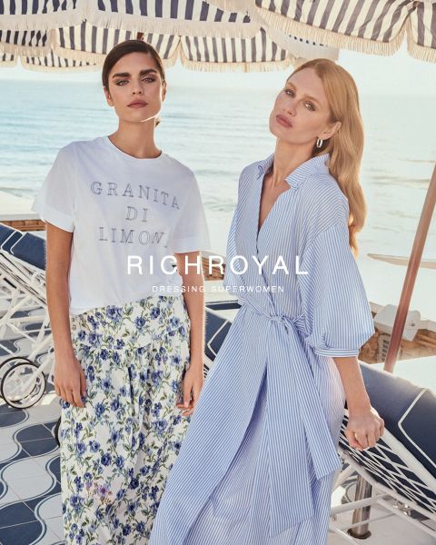 Zwei Frauen stehen mit gestreiften Sonnenschirmen auf einer Strandterrasse. Eine trägt ein „Granita di Limone“-Shirt und einen geblümten Rock. Die andere trägt ein blau-weiß gestreiftes Kleid. Beide wirken entspannt. Der Text lautet: „RICHROYAL: SUPERFRAUEN ANZIEHEN.“.