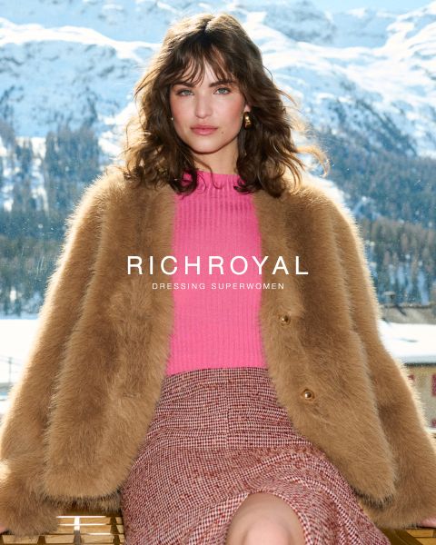 Eine Frau, die einen braunen Kunstpelzmantel, einen rosa Pullover und einen rot karierten Rock trägt, sitzt im Freien mit verschneiten Bergen im Hintergrund. Der Text "RICH&ROYAL DRESSING SUPERWOMEN" wird über dem Bild angezeigt.