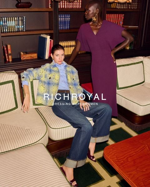 Zwei stilvolle Frauen posieren in einem mit Büchern ausgekleideten Raum; die eine sitzt in einer gelben Jacke und Jeans, die andere steht in einem lila Kleid. Der Text lautet: "Reich & Königlich.".