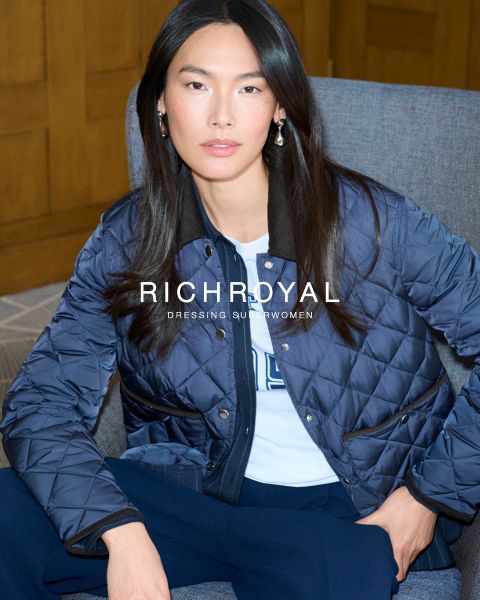 Eine Frau mit langen dunklen Haaren sitzt in einem grauen Stuhl und trägt eine blaue Steppjacke und marineblaue Hosen. Der Text auf dem Bild lautet "RICH&ROYAL DRESSING SUPERWOMEN". Sie blickt selbstbewusst in die Kamera.