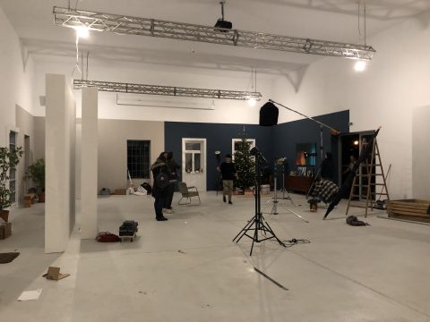 Ein geräumiger Raum, der als Filmset eingerichtet ist. Crewmitglieder stellen die Beleuchtungsausrüstung und ein Mikrofon auf einem Ständer ein. Die Wände sind in Dunkelblau und Weiß gestrichen. Im Raum sind Topfpflanzen, eine Leiter und verschiedene Requisiten verteilt.