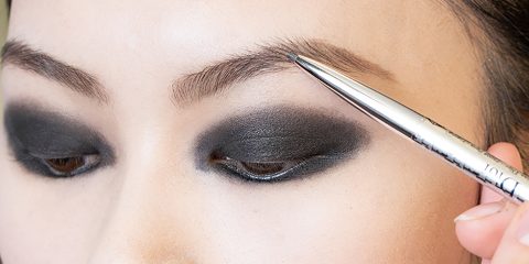 Eine Person schminkt sich mit einem silbernen Augenbrauenstift über einem dunklen Smokey-Eye-Look. Der Fokus liegt auf der Augenpartie mit kräftigem schwarzen Lidschatten und sauber geformten Augenbrauen.