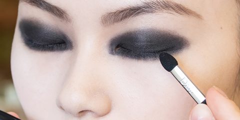 Nahaufnahme einer Person, die mit einem kleinen Applikator dunklen Lidschatten auf das Augenlid aufträgt und so einen kräftigen, rauchigen Augen-Make-up-Look kreiert. Der Fokus liegt auf der Augenpartie, die die intensive, verblendete schwarze Farbe zur Geltung bringt.