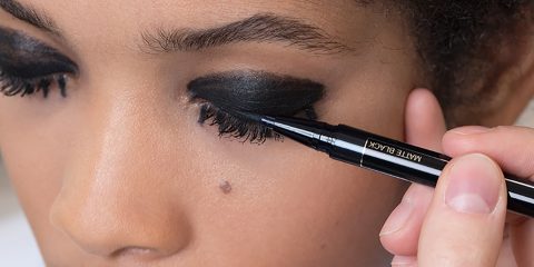 Eine Nahaufnahme einer Person, die schwarzen Eyeliner auf ihr Augenlid aufträgt. Ihr Auge ist geschlossen und der Eyeliner erzeugt einen kräftigen, dramatischen Blick. Die Hand, die den Eyeliner-Stift hält, ist ruhig und konzentriert sich auf das Auftragen.
