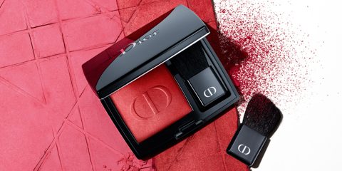 Ein Dior-Make-up-Kompakt mit rotem Rouge ist geöffnet und gibt den Blick auf zwei Pinsel frei. Der Hintergrund weist eine strukturierte Oberfläche auf, die teilweise mit rotem Puder bedeckt ist.