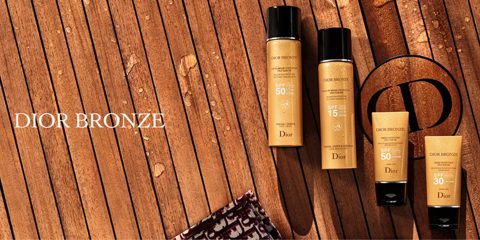 Eine Kollektion von Dior Bronze Sonnenschutzprodukten mit Lichtschutzfaktor 30, 50 und 15 wird auf einer Holzoberfläche präsentiert. In der Ecke ist teilweise ein Handtuch mit gemustertem Rand sichtbar. Das Dior-Logo und der Text „DIOR BRONZE“ befinden sich auf der linken Seite.
