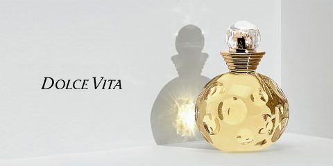 Eine runde, verzierte Parfümflasche mit einem goldenen und transparenten Design steht auf einer reflektierenden Oberfläche. Licht wirft einen deutlichen Schatten. Der Text „Dolce Vita“ steht auf der linken Seite des Bildes.