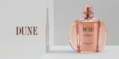Eine Flasche Dune Eau de Toilette von Christian Dior wird auf einer weißen Oberfläche und einem weißen Hintergrund präsentiert. Die Flasche reflektiert einen weichen Schatten und der Markenname „DUNE“ ist deutlich daneben zu sehen. Das Parfüm hat einen roségoldenen Farbton.