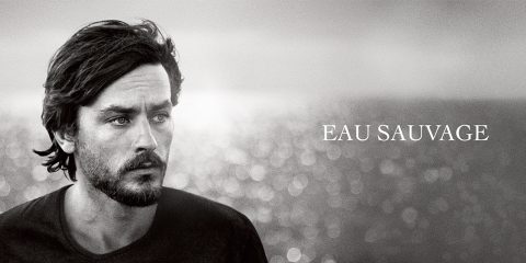 Schwarzweißbild eines Mannes mit Bart, der zur Seite blickt, vor einem verschwommenen Meereshintergrund. Rechts wird der Text „EAU SAUVAGE“ angezeigt.