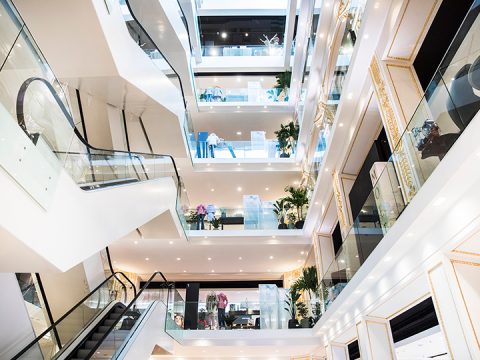 Modernes mehrstöckiges Einkaufszentrum mit Rolltreppen, Glasgeländern und mehreren Ebenen mit verschiedenen Ladenfronten. Das Design zeichnet sich durch helle Beleuchtung, Topfpflanzen und offene Räume aus, die eine luftige und moderne Atmosphäre schaffen.
