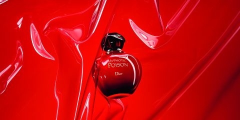 Eine Flasche Parfüm Dior Hypnotic Poison steht vor einem leuchtend roten Hintergrund mit wirbelnden Texturen, was das luxuriöse und verführerische Design hervorhebt.