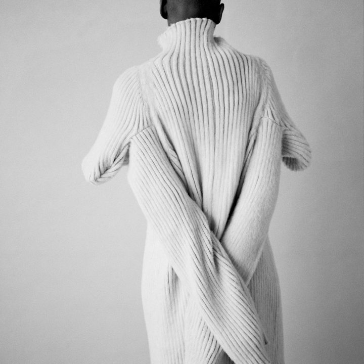 Von hinten ist eine Person mit rasiertem Kopf zu sehen, die einen übergroßen Rippenstrickpullover mit überlangen, herunterhängenden Ärmeln trägt. Der Hintergrund ist in einem schlichten, hellgrauen Farbton gehalten.