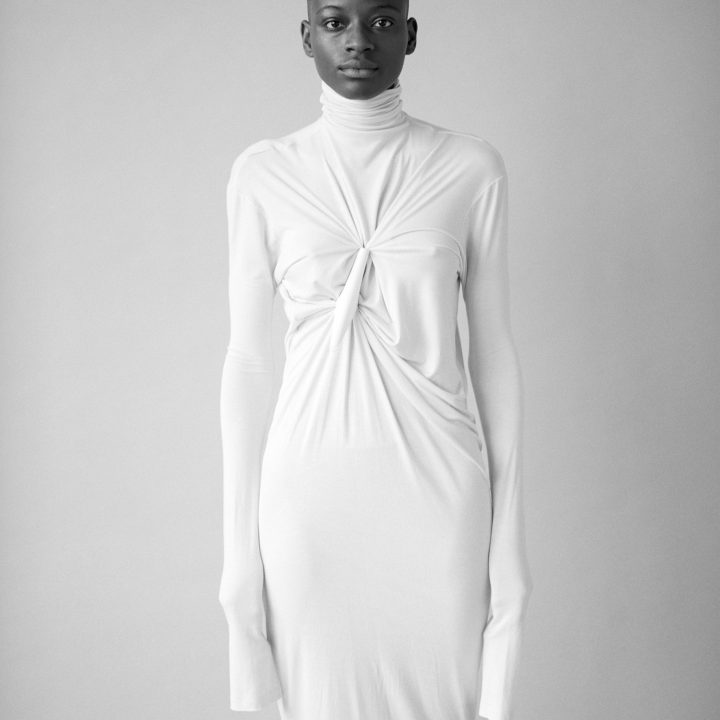Eine Person steht vor einem neutralen Hintergrund und trägt ein langärmeliges, weißes Kleid mit hohem Kragen und Knotendetail auf der Brust. Das Kleid reicht bis zur Mitte der Wade. Der Gesichtsausdruck der Person ist ruhig und direkt.