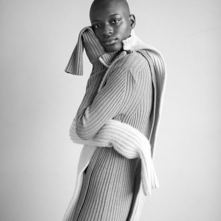 Eine Person posiert selbstbewusst in einem gemütlichen, langen Rippenstrickpullover mit passendem Schal. Das Bild ist schwarzweiß und hebt die Textur des Outfits hervor. Der Hintergrund ist einfach und neutral und lenkt den Fokus auf die Person.