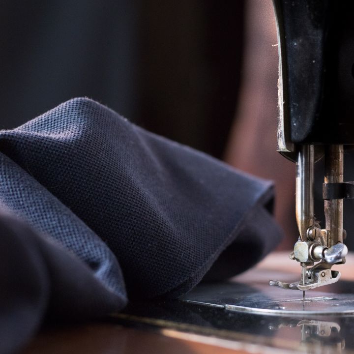 Nahaufnahme einer Nähmaschine, die dunklen Stoff näht. Der Fokus liegt auf Nadel und Nähfuß, wodurch die Textur des Stoffes und die Metallteile der Maschine hervorgehoben werden. Der Hintergrund ist zur Hervorhebung unscharf.