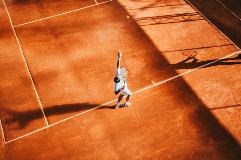 Ein Tennisspieler bereitet sich auf einem sonnenbeschienenen Sandplatz auf seinen Aufschlag vor. Der Schatten des Spielers und seines Schlägers ist auf der Oberfläche des Platzes sichtbar und unterstreicht die sportliche Bewegung. Die Szene fängt die dynamische Energie des Sports ein.