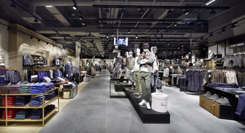 Ein modernes Interieur eines Bekleidungsgeschäfts mit Schaufensterpuppen, die lässige Outfits präsentieren. Regale und Ständer sind mit ordentlich sortierter Kleidung gefüllt und das Geschäft verfügt über ein minimalistisches Design mit Grautönen und Deckenbeleuchtung.