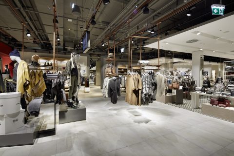 Ein geräumiges Bekleidungsgeschäft mit modernem, industriellem Design. Schaufensterpuppen präsentieren verschiedene Outfits, umgeben von Kleiderständern. Es herrschen neutrale Töne vor, reichlich Beleuchtung und hohe Decken schaffen eine offene, einladende Atmosphäre.