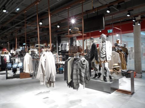 Ein modernes Bekleidungsgeschäft mit Regalen voller weißer und grauer Kleidungsstücke, Hüte und Accessoires. Schaufensterpuppen tragen legere Kleidung. Das Geschäft ist großzügig gestaltet, mit Deckenbeleuchtung und freiliegender Decke.
