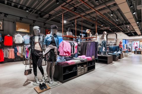 Ein modernes Interieur eines Bekleidungsgeschäfts mit Schaufensterpuppen in legerer Kleidung, umgeben von mehreren Ständern mit farbenfroher Kleidung. Das Geschäft ist hell beleuchtet und in den Regalen liegen ordentlich gefaltete Kleidungsstücke.