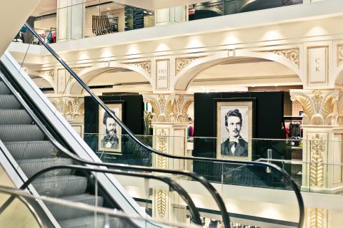 Das Interieur eines luxuriösen Kaufhauses mit kunstvollem Design. Rolltreppen durchziehen die Szene und führen zu verschiedenen Ebenen. Große gerahmte Porträts unbekannter Figuren schmücken die Wände. Im Hintergrund sieht man Käufer, die stöbern.