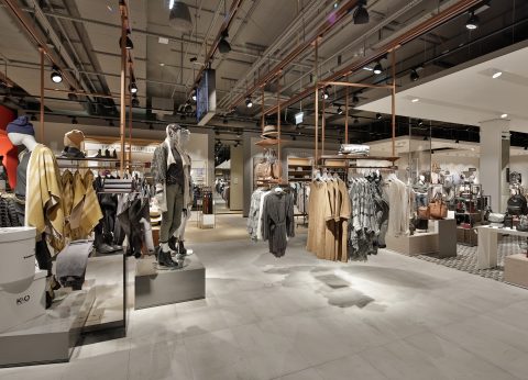 Ein modernes Interieur eines Bekleidungsgeschäfts mit Schaufensterpuppen, die eine Vielzahl von Outfits präsentieren. Der Raum verfügt über Regale mit ordentlich angeordneter Kleidung und Accessoires sowie warmes Licht, das eine einladende Atmosphäre schafft. Der Boden ist hell und die Decke hat freiliegende Armaturen.