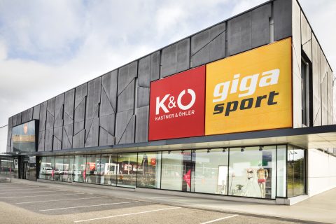 Ein modernes Gebäude mit großen Glasfenstern und geometrischen Paneelen zeigt die Logos von „K&Ö Kastner & Öhler“ und „giga sport“. Leuchtend orange und rote Schilder bilden einen Kontrast zum grauen Äußeren. Im Vordergrund ist ein Parkplatz zu sehen.