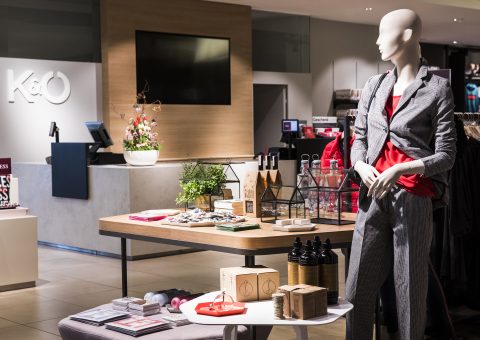 Schaufensterpuppe in einem stilvollen grauen Outfit mit rotem Oberteil, ausgestellt in einem modernen Einzelhandelsgeschäft. Der Tisch daneben ist mit verschiedenen Produkten und Dekorationsartikeln geschmückt. Im Hintergrund sind eine Kasse, ein Fernsehbildschirm und ein „K&O“-Logo zu sehen.