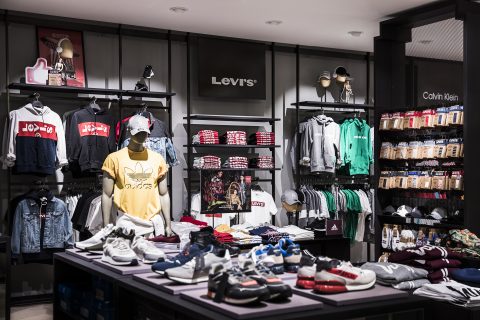 Schaufenster eines Bekleidungsgeschäfts mit einer Auswahl an Kleidung und Schuhen. Schaufensterpuppen tragen Marken-Kapuzenpullover und T-Shirts unter Levi's-Schildern. Auf Regalen liegen gefaltete Hemden, Mützen und Accessoires, während auf einem zentralen Tisch Turnschuhe ausgestellt sind.