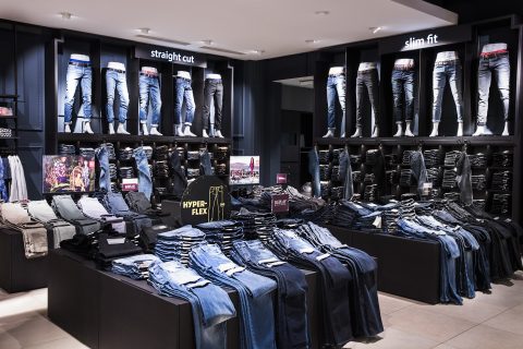 Eine Schaufensterpuppe präsentiert verschiedene Jeans in einem Einzelhandelsgeschäft. Schaufensterpuppen präsentieren gerade geschnittene und schmal geschnittene Modelle über Regalen voller ordentlich gefalteter Jeans in verschiedenen Farbtönen. Schilder heben Merkmale wie „HYPERFLEX“ hervor.