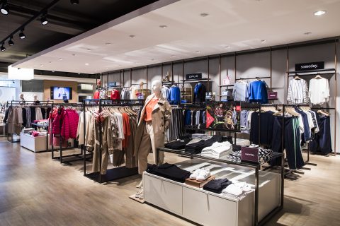 Ein modernes Interieur eines Bekleidungsgeschäfts mit ordentlich angeordneten Regalen voller bunter Hemden, Jacken und Mäntel. Im Vordergrund steht eine Schaufensterpuppe in einem Trenchcoat. Das Geschäft hat einen warmen Holzboden und helle Deckenbeleuchtung.