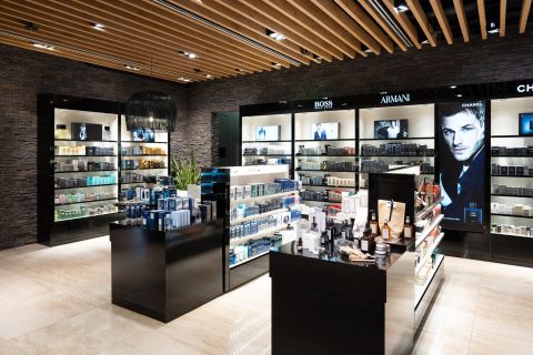 Ein modernes Ladeninterieur mit Regalen, in denen verschiedene Luxusdüfte und Schönheitsprodukte ausgestellt sind. Der Laden hat Holzakzente an der Decke, elegante schwarze Regale und leuchtende Schilder für Marken wie Boss, Armani und Chanel.
