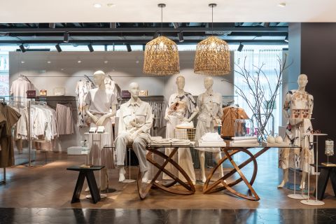 Ein modernes Schaufenster in einem Bekleidungsgeschäft mit Schaufensterpuppen in neutralen Outfits. Die Schaufensterpuppen sind um einen Holztisch mit Dekorationsgegenständen angeordnet. Über ihnen hängen geflochtene Hängelampen und an den Wänden hängen Kleiderständer.