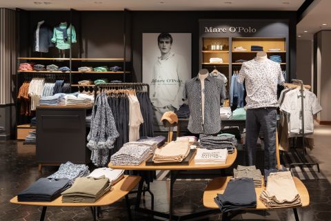 Eine Auslage in einem Bekleidungsgeschäft mit ordentlich gefalteten Hemden und Hosen auf Tischen. Schaufensterpuppen tragen gemusterte Hemden und dunkle Hosen. Auf Regalen im Hintergrund stehen verschiedene bunte Hemden. An der Wand hängt ein großes Poster einer Person.
