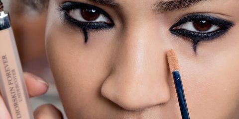 Eine Nahaufnahme des Gesichts einer Person mit dramatischem Augen-Make-up, mit dickem Eyeliner und gezeichneten Tränenlinien. Sie trägt mit einem kleinen Pinsel Concealer unter dem Auge auf. Auf der Concealer-Flasche steht „Diorskin Forever Undercover“.
