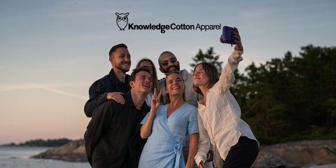Eine Gruppe von fünf Personen macht draußen in der Nähe von Wasser ein Selfie, mit Bäumen und einem Sonnenuntergang im Hintergrund. Sie lächeln und posieren verspielt, in legerer Kleidung. Über ihnen sind der Text „KnowledgeCotton Apparel“ und ein Eulenlogo zu sehen.