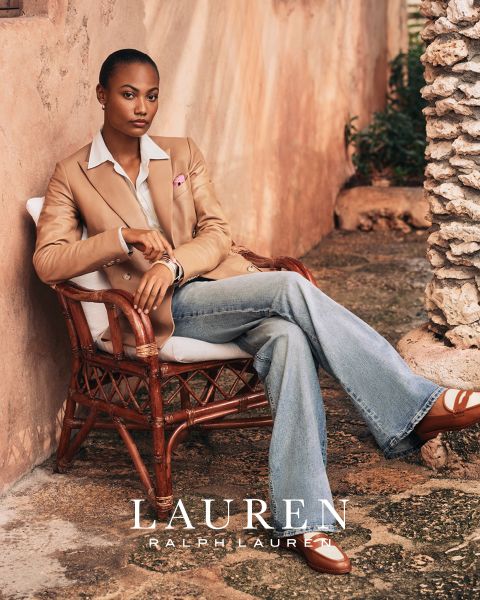 Eine Person sitzt im Freien in einem Korbstuhl vor einem rustikalen Hintergrund. Sie trägt einen hellbraunen Blazer, ein hellblaues Hemd und Schlagjeans mit braunen und weißen Schuhen. Der Text unten lautet „LAUREN RALPH LAUREN“.