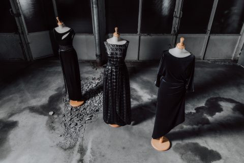 Drei Schaufensterpuppen präsentieren elegante schwarze Abendkleider. Jedes Kleid hat einen eigenen Stil und weist unterschiedliche Texturen und Schnitte auf. Sie stehen auf einem strukturierten Betonboden in einer schwach beleuchteten, industriellen Umgebung.