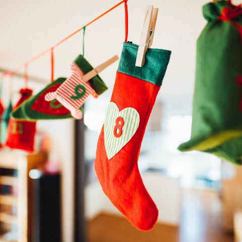 Eine Reihe bunter Weihnachtsstrümpfe und -säckchen ist mit Wäscheklammern an einer roten Schnur aufgehängt. Der mittlere Strumpf ist rot und grün, geschmückt mit einem weißen Herz und der Zahl 8. Der Hintergrund ist leicht verschwommen.