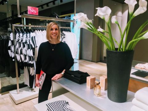Eine Person in einem Geschäft steht neben einem Ausstellungstisch mit einer großen Vase mit weißen Blumen und gefalteter Kleidung. Hinter ihr steht ein Kleiderständer mit einfarbigen Kleidungsstücken. Sie lächelt und hält einen roten Ordner in der Hand.